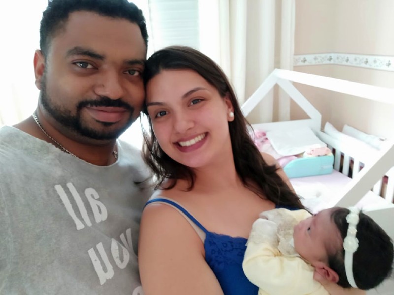 Foto de Gislaine e Melissa com o pai no quarto da bebê. Gislaine e o marido olham para a foto e sorriem. A mãe segura a bebê no colo, que está com uma faixa branca na cabeça e macacão amarelo. Do lado direito aparece parte do berço de Melissa